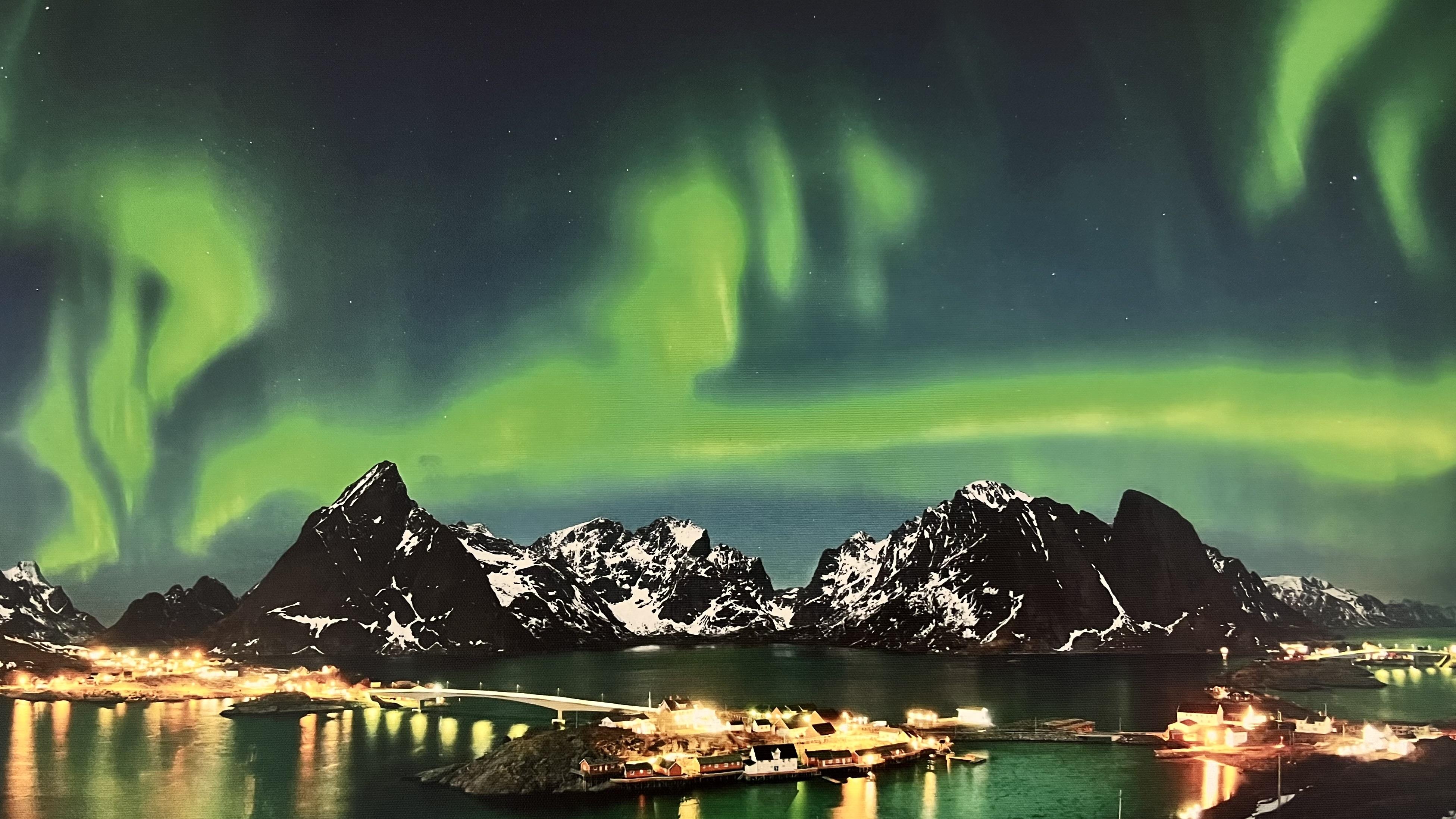 Grüne Polarlichter über schwarzen Bergen, die sich auf einer Wasseroberfläche im Vordergrund spiegeln.